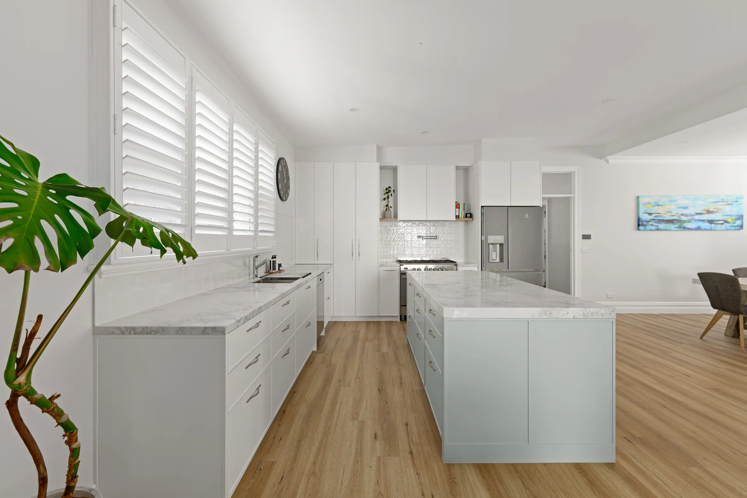 Kitchen with white flat-panel base drawers, marble-look stone benchtops, tall white pantry cupboards and a long pale sage island on timber-look flooring.