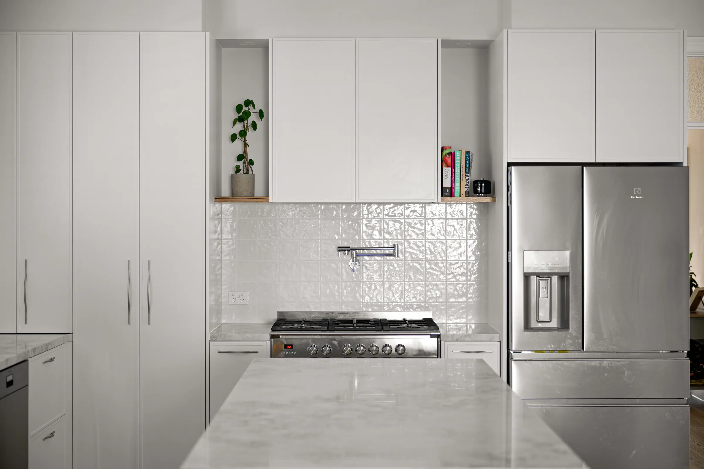 Cooktop wall with white overhead cupboards, open niches on timber shelves, textured gloss white tiles, pot filler and tall pantry doors.