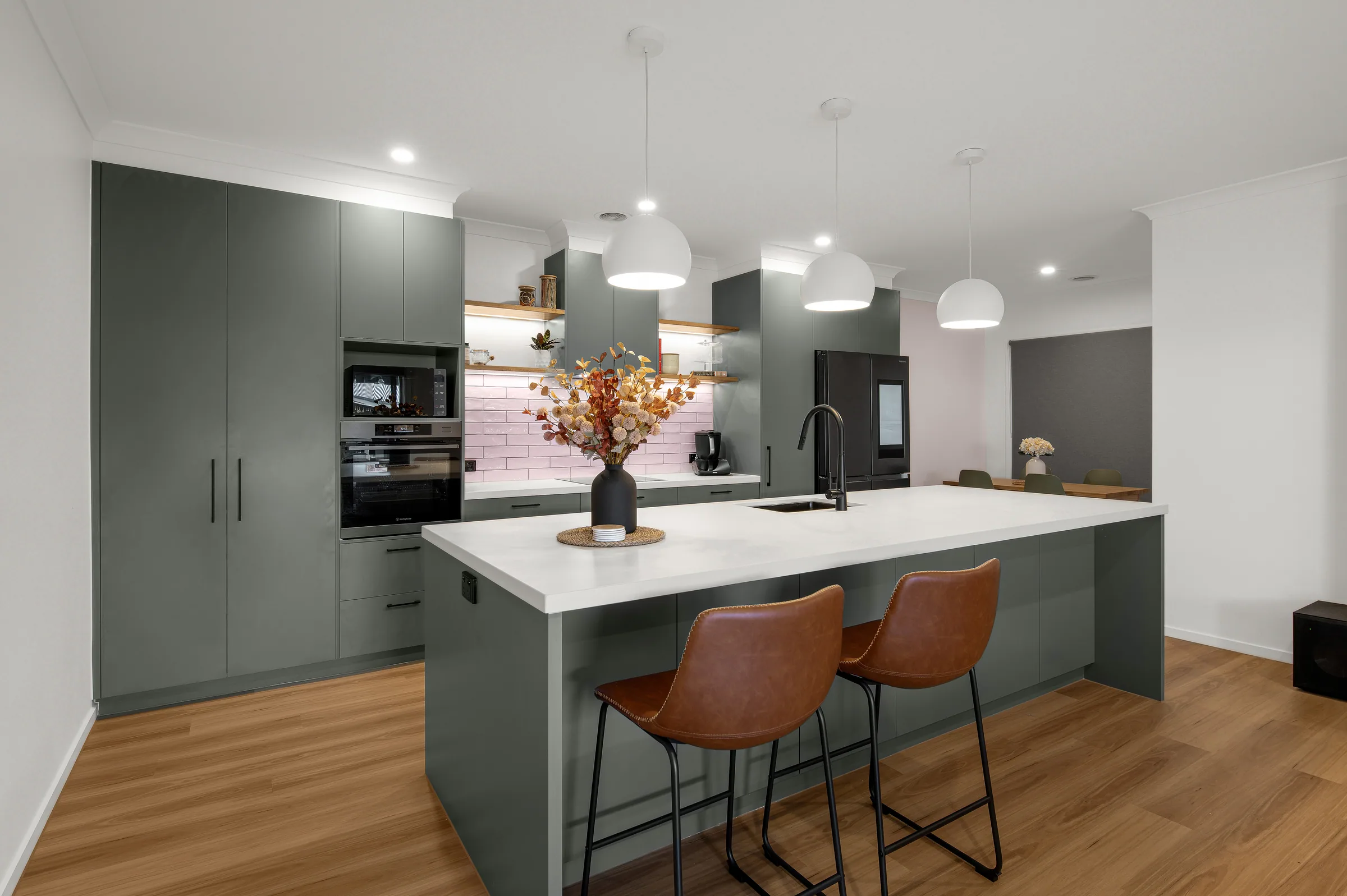 Sage green kitchen with full-height cabinetry, black ovens, white stone island bench, three white pendant lights and tan leather stools on timber floor.