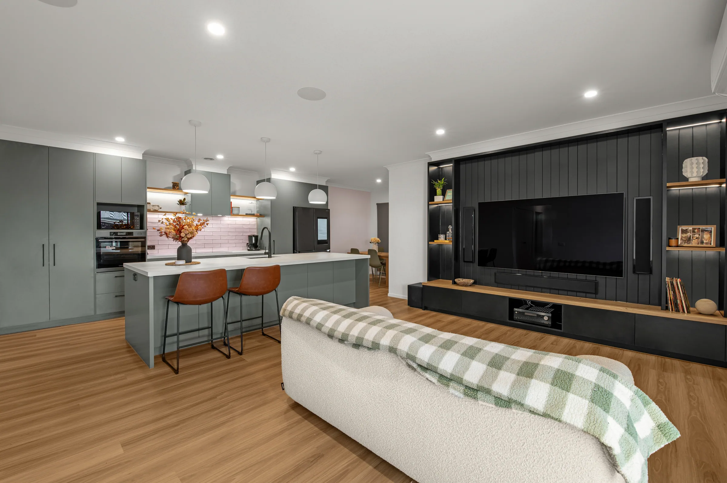 Open-plan room showing the sage green kitchen and island on the left and the black vertical-board media wall with timber bench on the right.