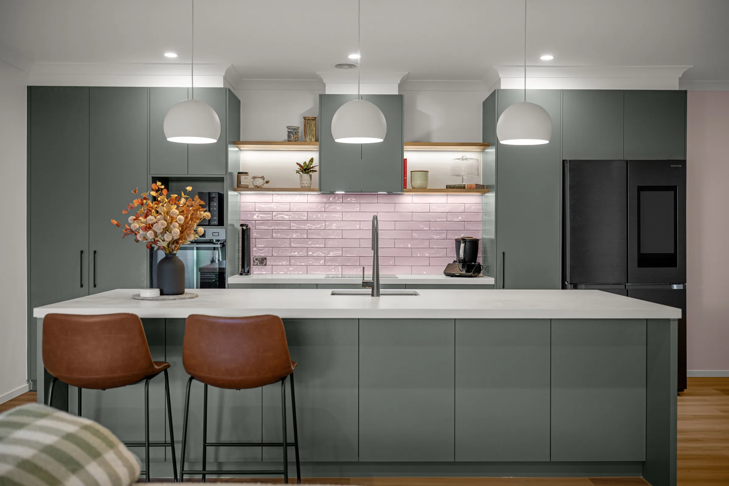Front-on view of the kitchen: green cabinetry either side of a pink tiled splashback, timber floating shelves, pendants and a long green island with white benchtop.