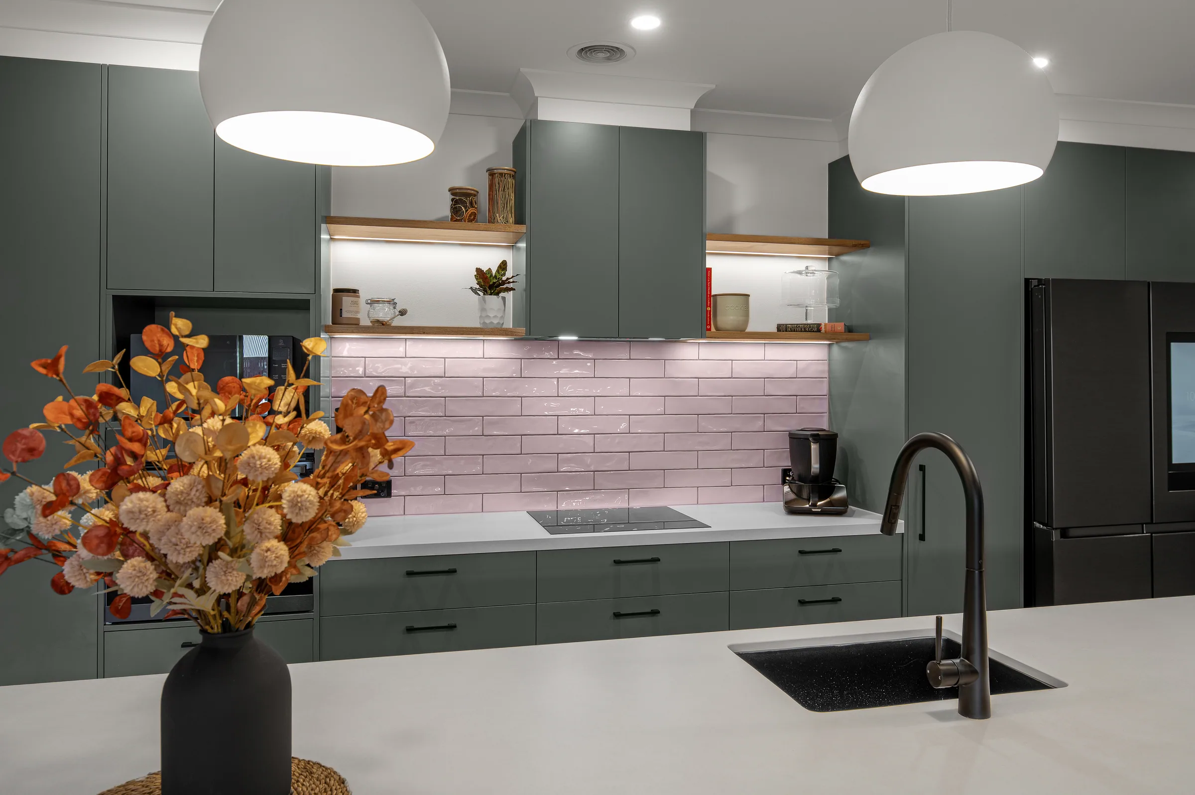 Cooking run with pink gloss subway tiles, lit timber floating shelves, green overhead and base cabinets, black induction cooktop and black tapware.