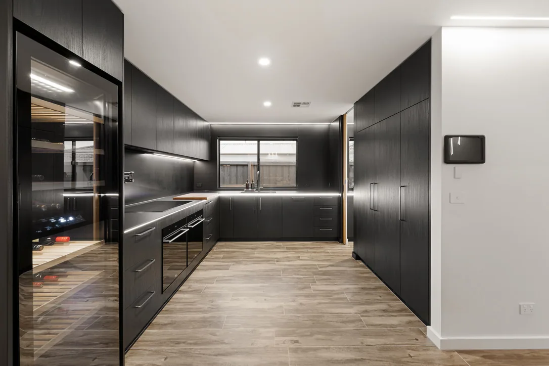 Wide view of a black timber-grain kitchen with a glass-fronted wine cabinet, tall cupboard bank, strip lighting under overheads and pale timber-look floor.
