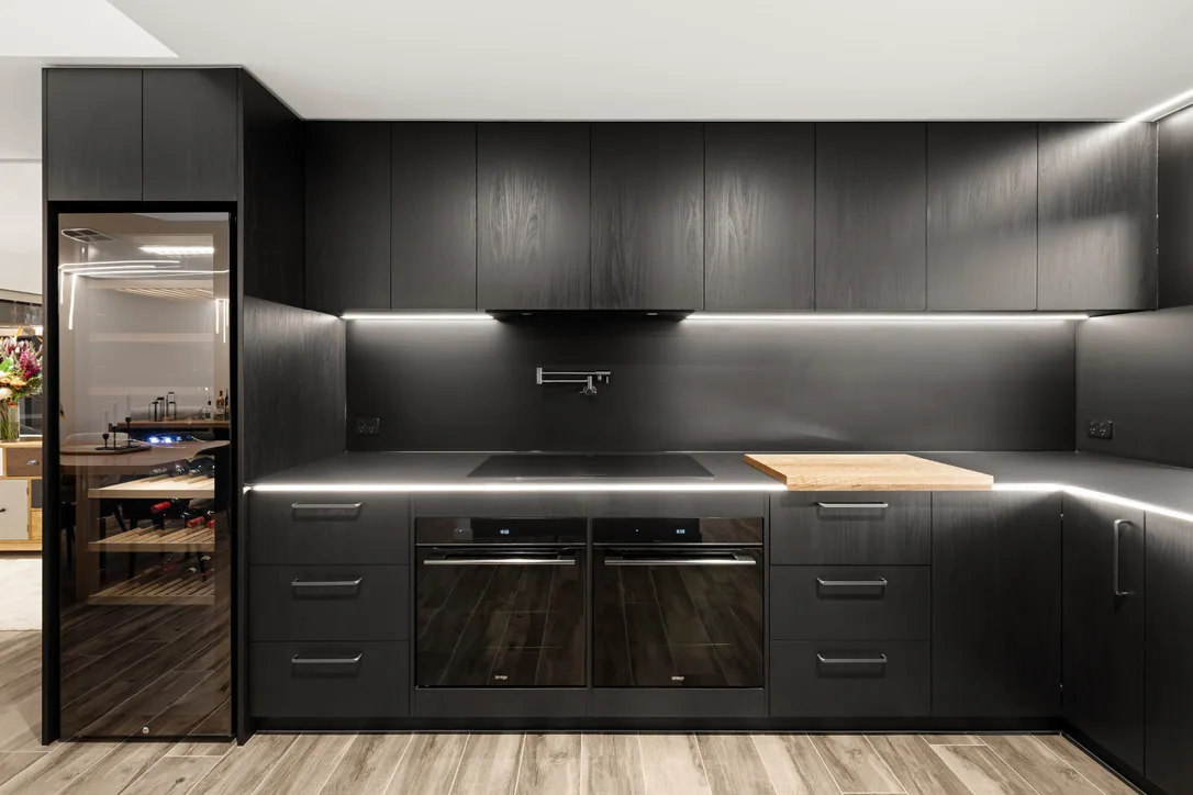 Straight-on elevation of black kitchen cabinetry with twin black ovens, induction cooktop, dark stone benchtop, under-cabinet lighting and a timber chopping board.
