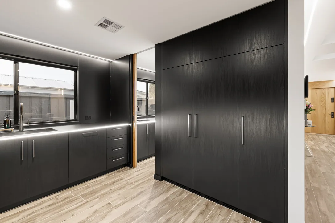 Black cabinetry along a window sink run beside a floor-to-ceiling bank of black doors with slim steel handles on pale timber-look flooring.