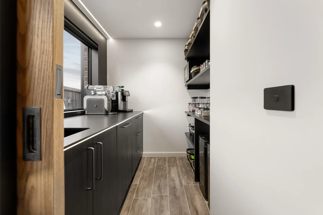 Butler's pantry with black base cabinets, dark benchtop, open black shelving and a timber sliding door with a recessed black pull.