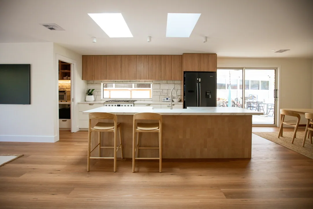 Front view of a kitchen with timber-veneer overhead cabinets, a tiled island with white stone benchtop, two timber stools and skylights above.