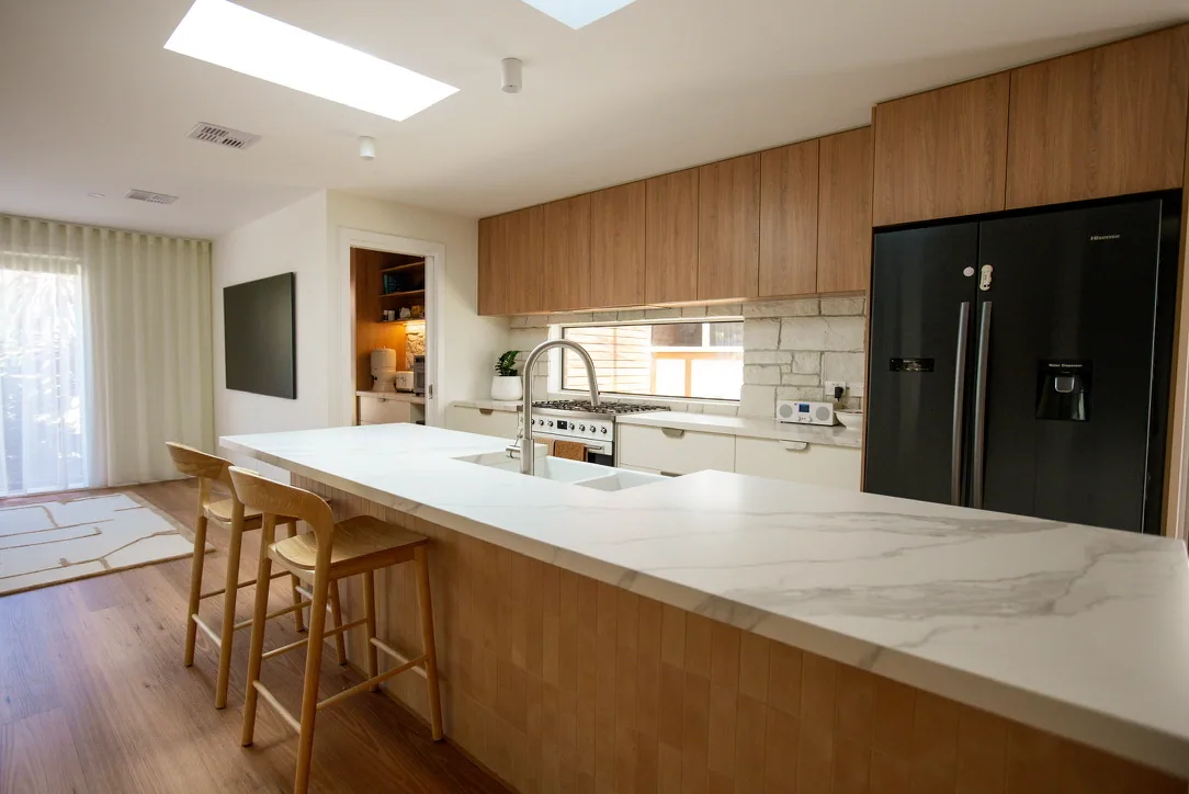 Angled view along the island benchtop toward timber overhead cabinets, a split-face stone splashback, window and black fridge.