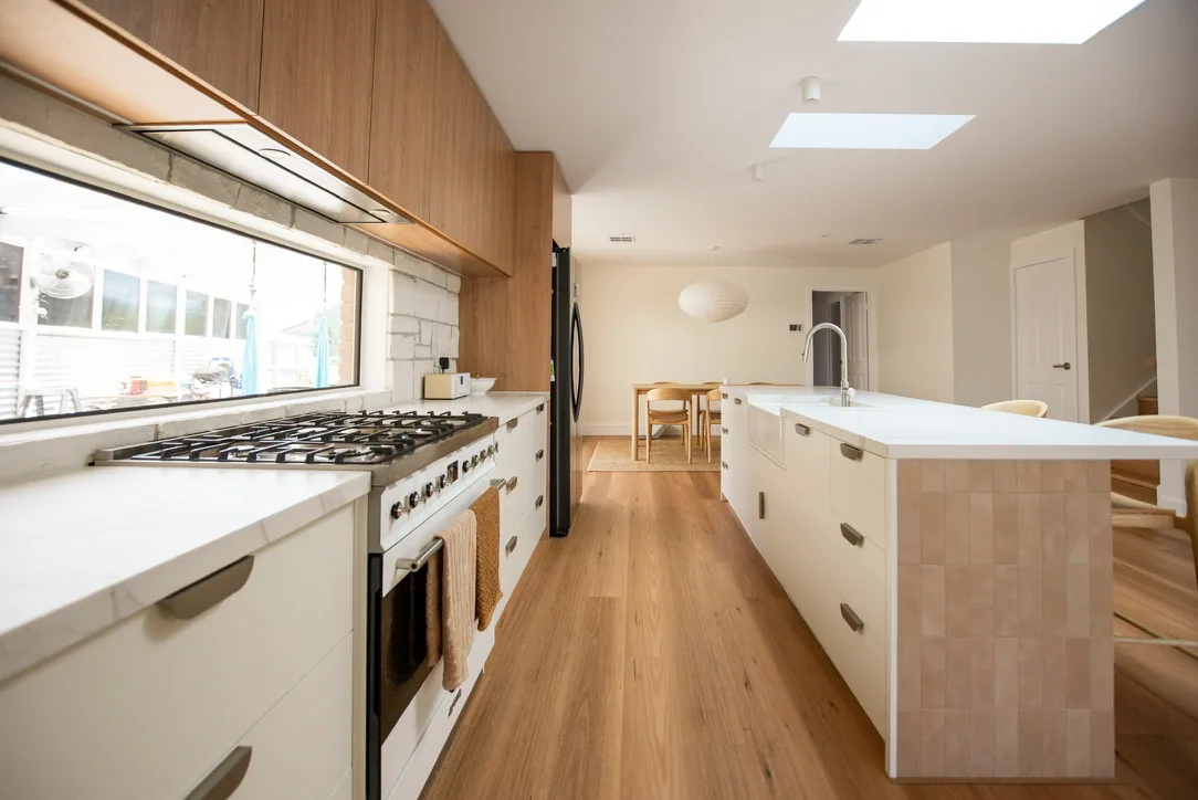Kitchen run with white drawer fronts, brushed pull handles, freestanding cooker, window splashback and timber overheads, island to the right.