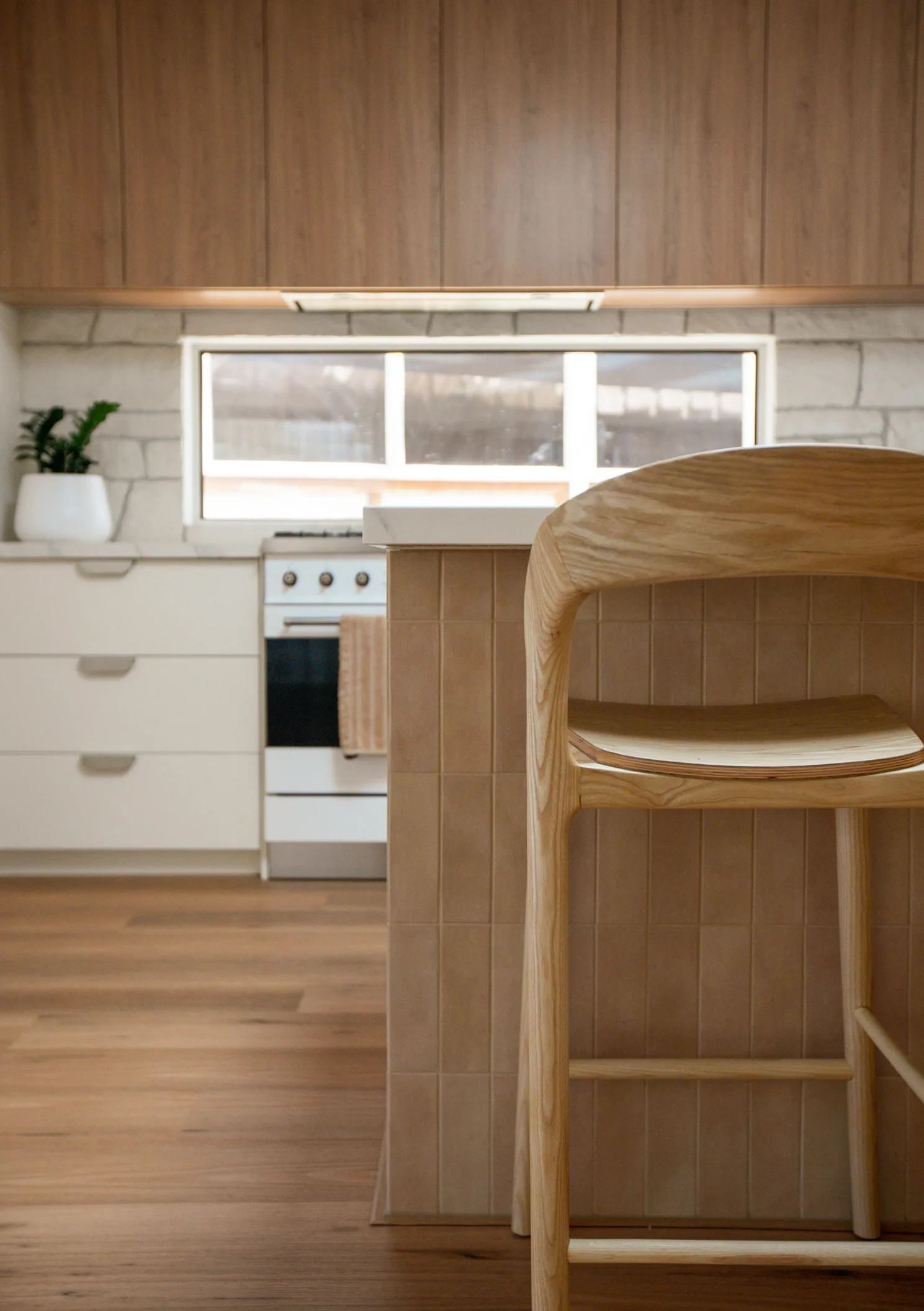 Close view of a timber stool against the pale tiled island end, with white drawers, cooker and stone splashback behind.