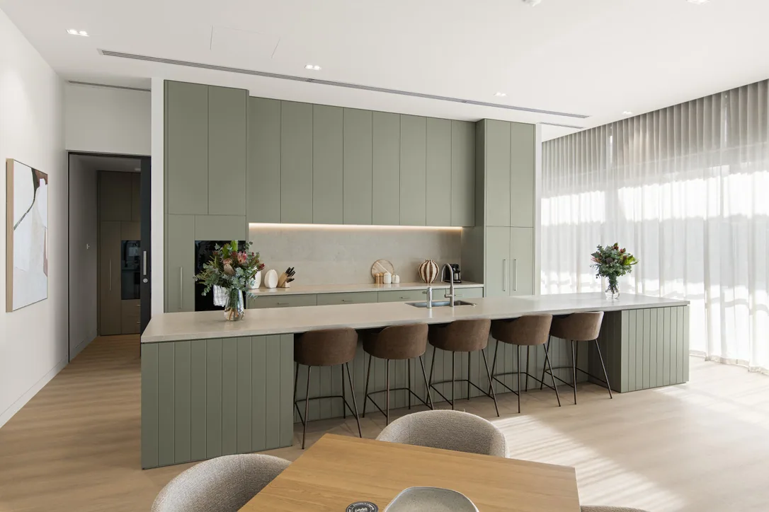 Sage-green kitchen with full-height flat-panel cabinetry, a lit stone splashback and a long vertical-batten island with a pale stone top.