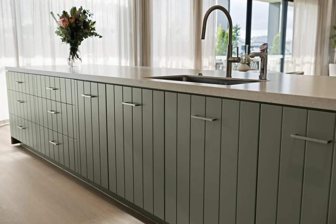 Close view of the island's sage-green vertical-batten drawer and door fronts with slim brushed metal handles and an undermount sink.