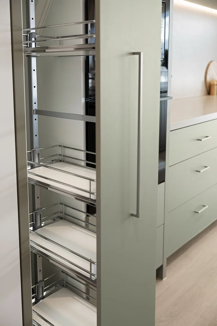 Tall sage-green pantry unit pulled open to show chrome wire basket shelves, with a long bar handle on the door.