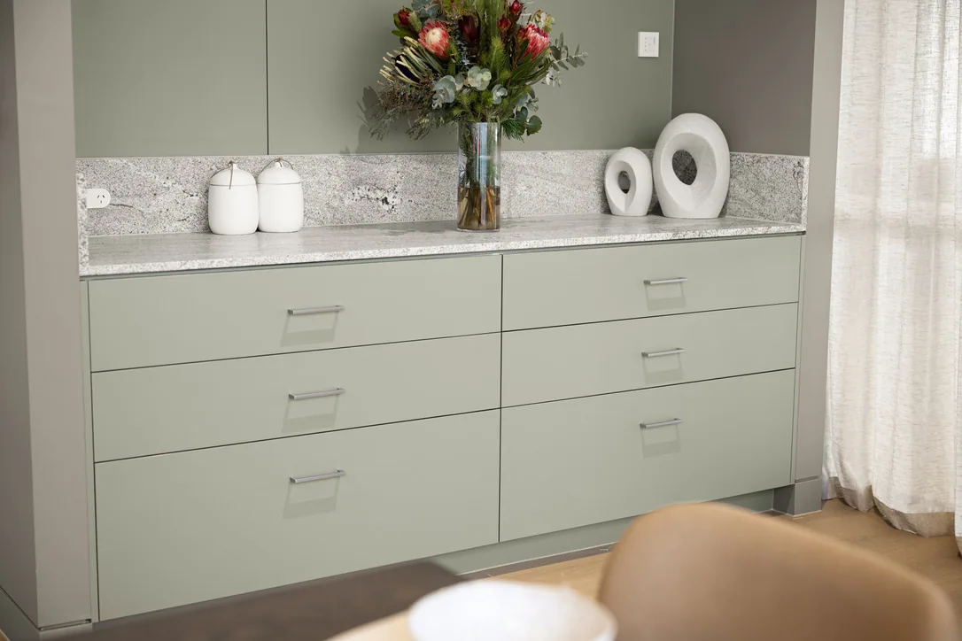 Sage green drawer bank with slim bar handles under a grey veined stone bench, with a vase of flowers and ceramics on top.
