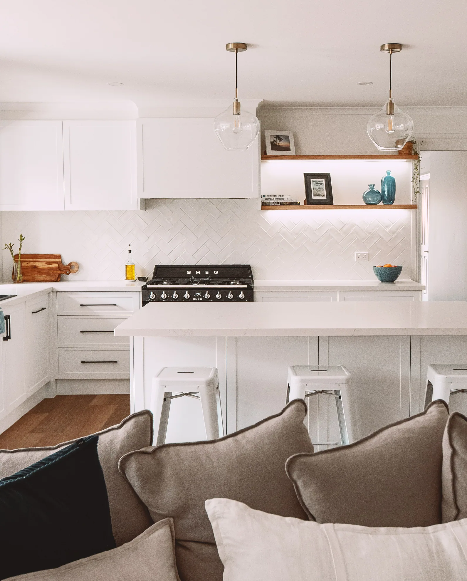 White shaker kitchen with a panelled island, a cabinetry canopy over a black SMEG cooker, herringbone tiles and two lit timber floating shelves.