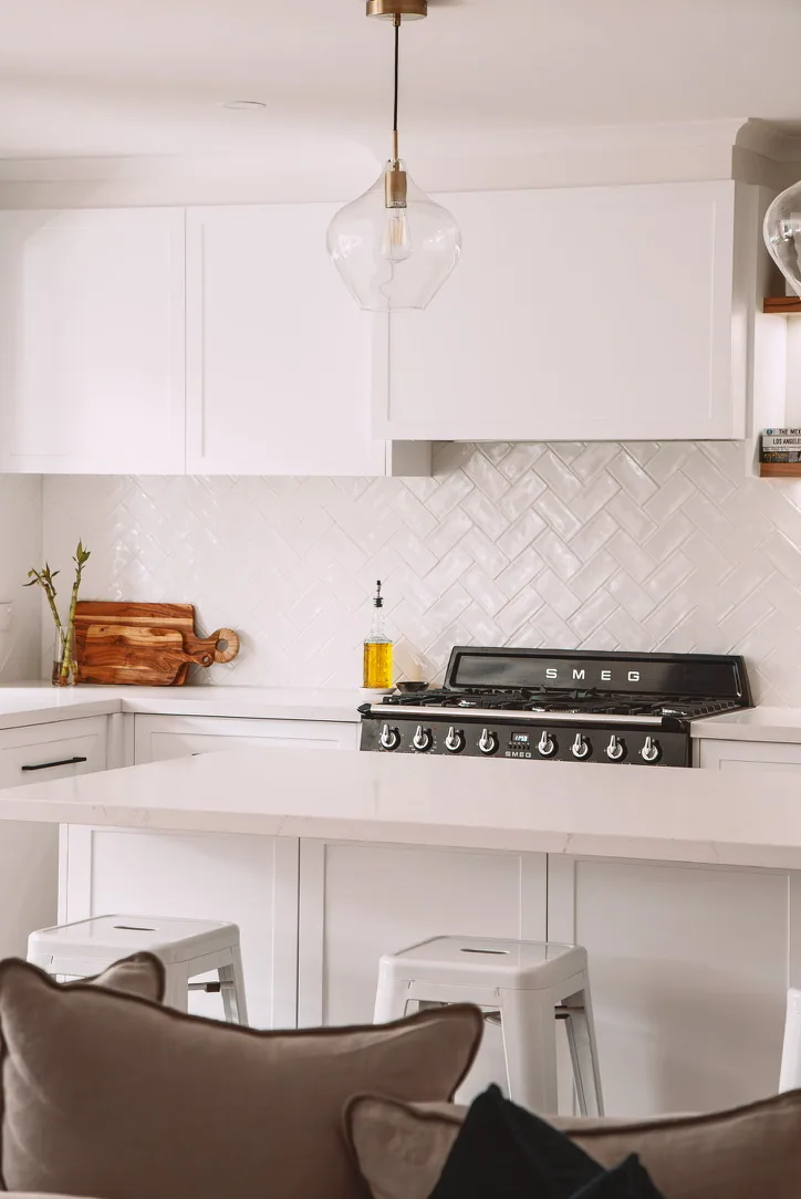White shaker wall cabinets and rangehood canopy above glossy herringbone tiles and a black SMEG cooker, with a panelled island and white stools in front.