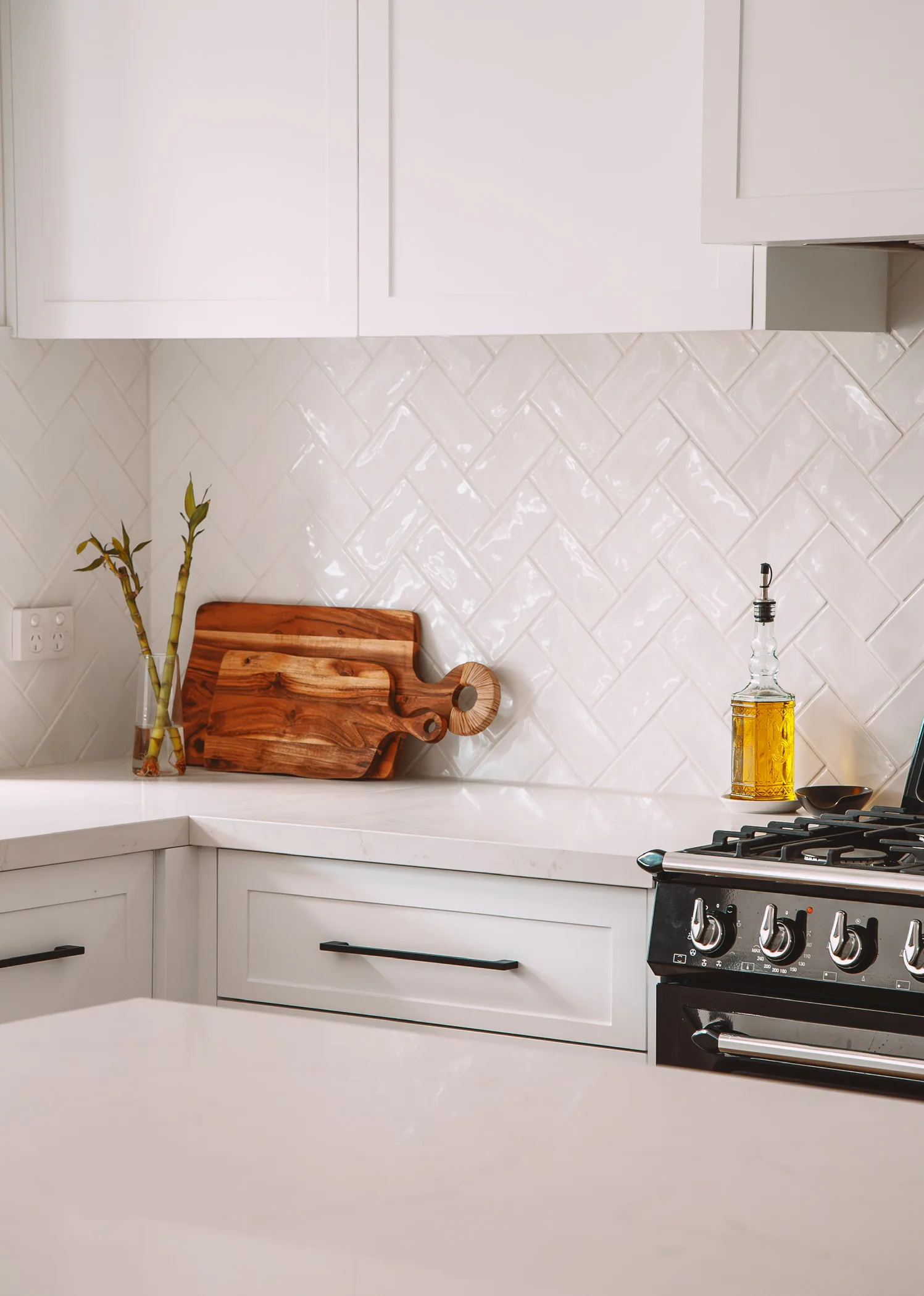 Corner of white shaker drawers and wall cabinets with slim black bar handles, a white stone-look benchtop and glossy herringbone tiles.
