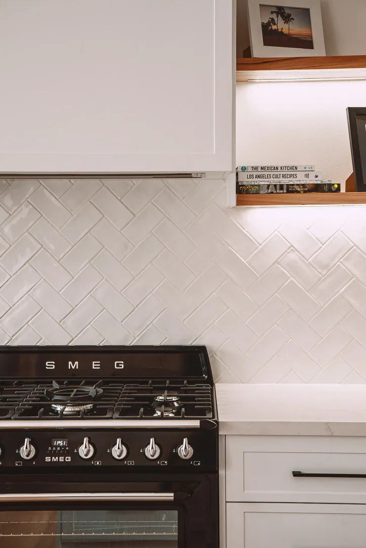White shaker rangehood canopy beside two timber floating shelves with an LED strip under the upper shelf, above a black SMEG cooker, herringbone tiles and a white drawer with a black bar handle.
