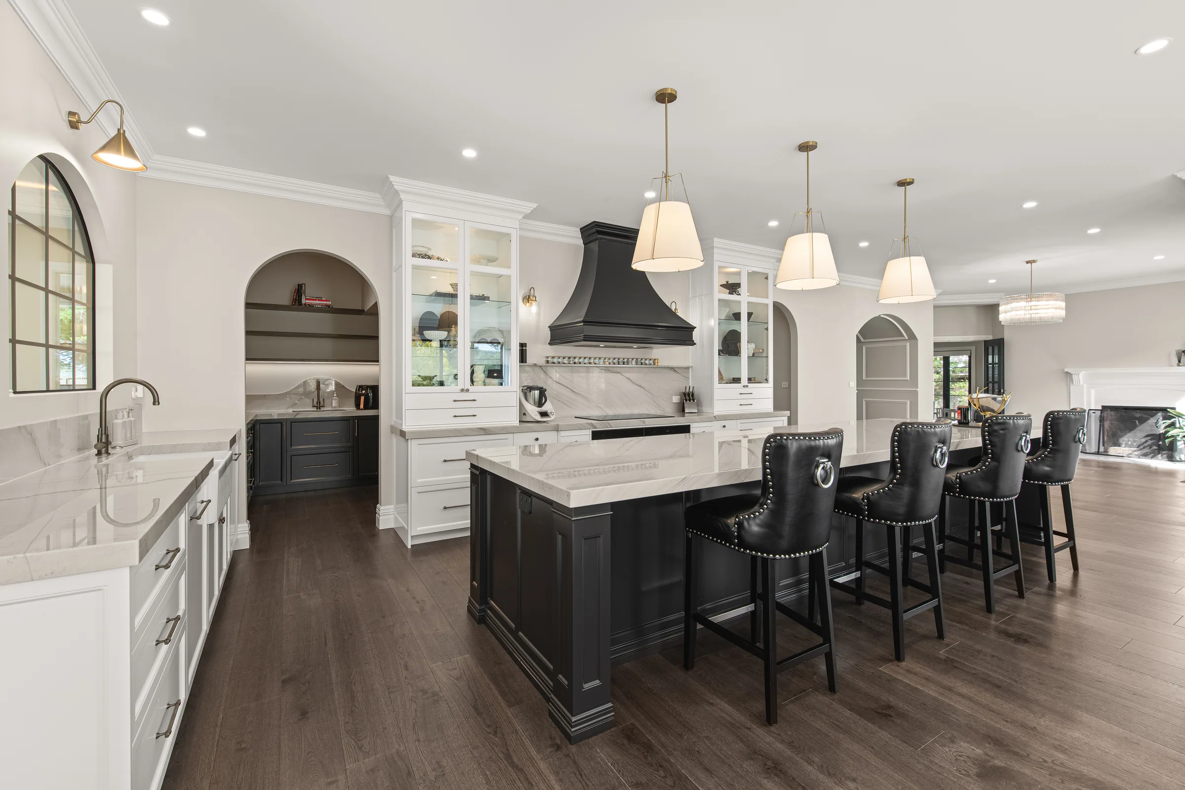 Wide view of white wall cabinetry, a black panelled island with leather stools, and an arch into the pantry.