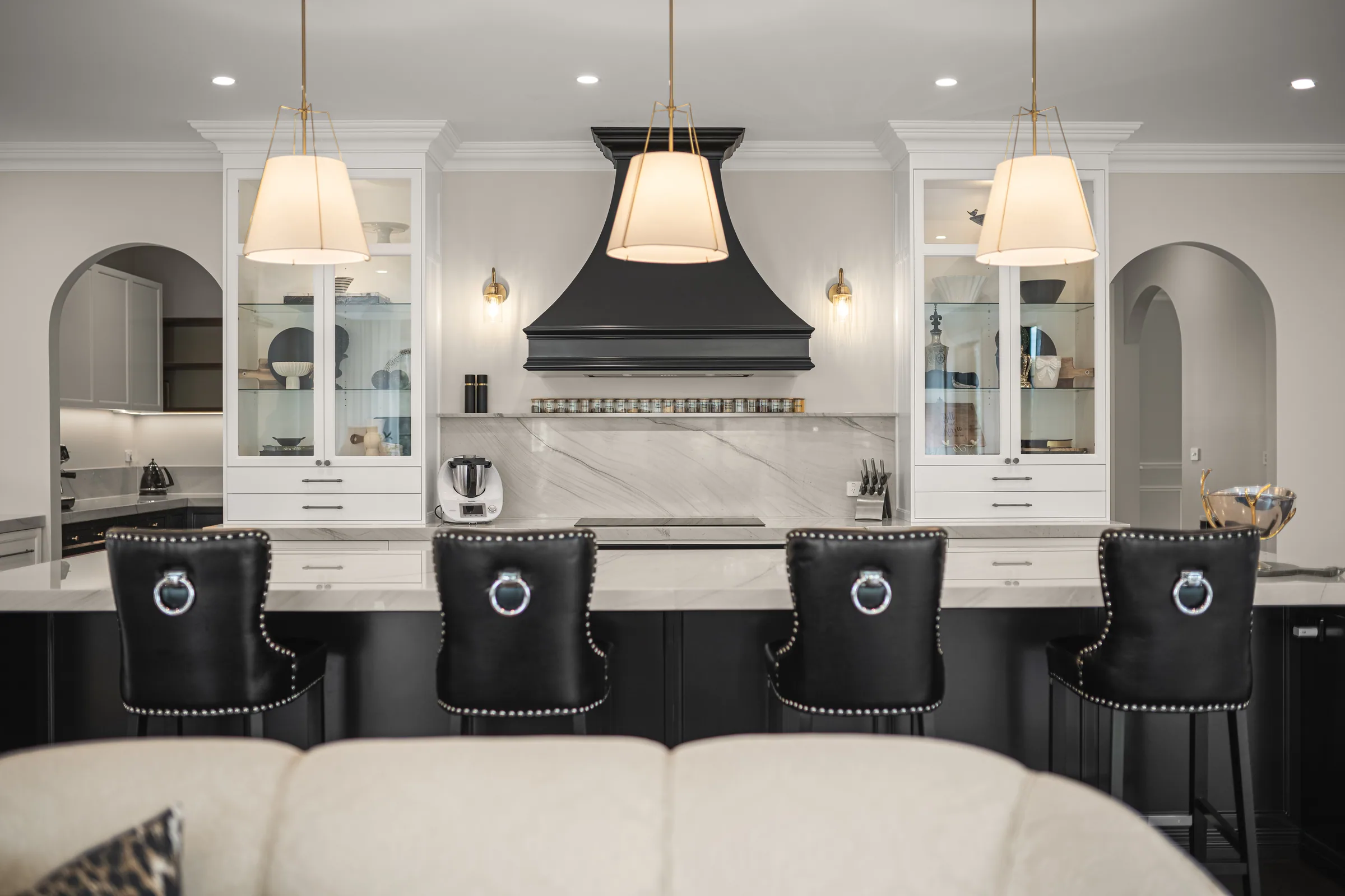 Black curved range hood between two glazed white display cabinets, with three linen pendants above a stone-topped black island.