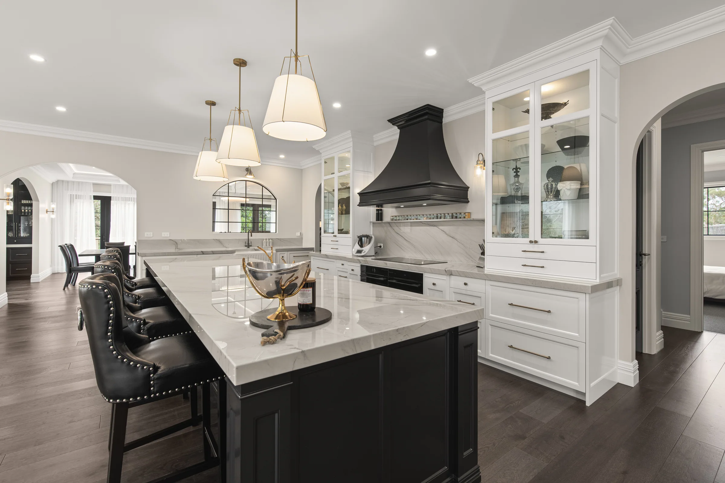 Black island with pale veined stone top and studded leather stools, white cabinetry and black range hood behind.