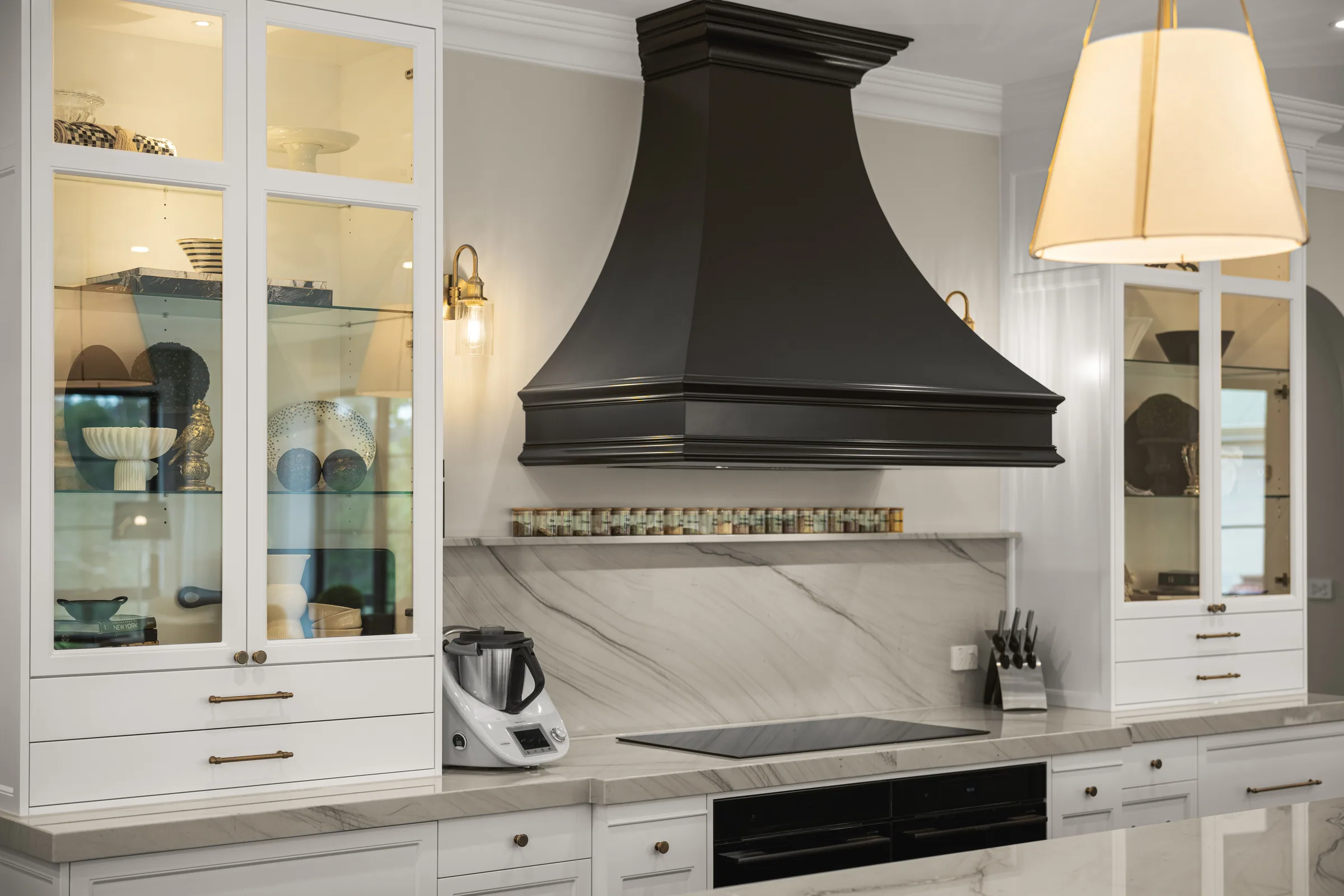 Black moulded range hood over a veined stone splashback and spice shelf, flanked by lit glass-fronted white cabinets.