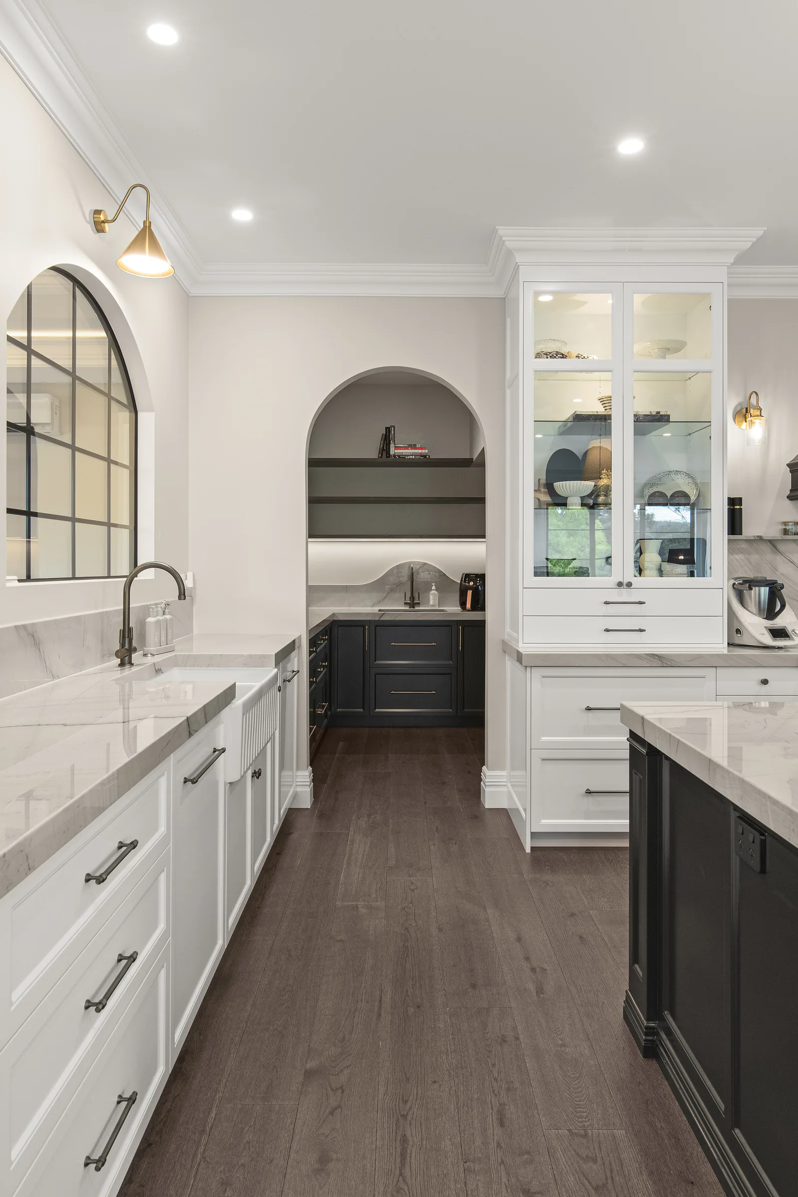 White shaker drawers with brass pulls beside a fluted farmhouse sink, looking through an arch to the dark pantry.