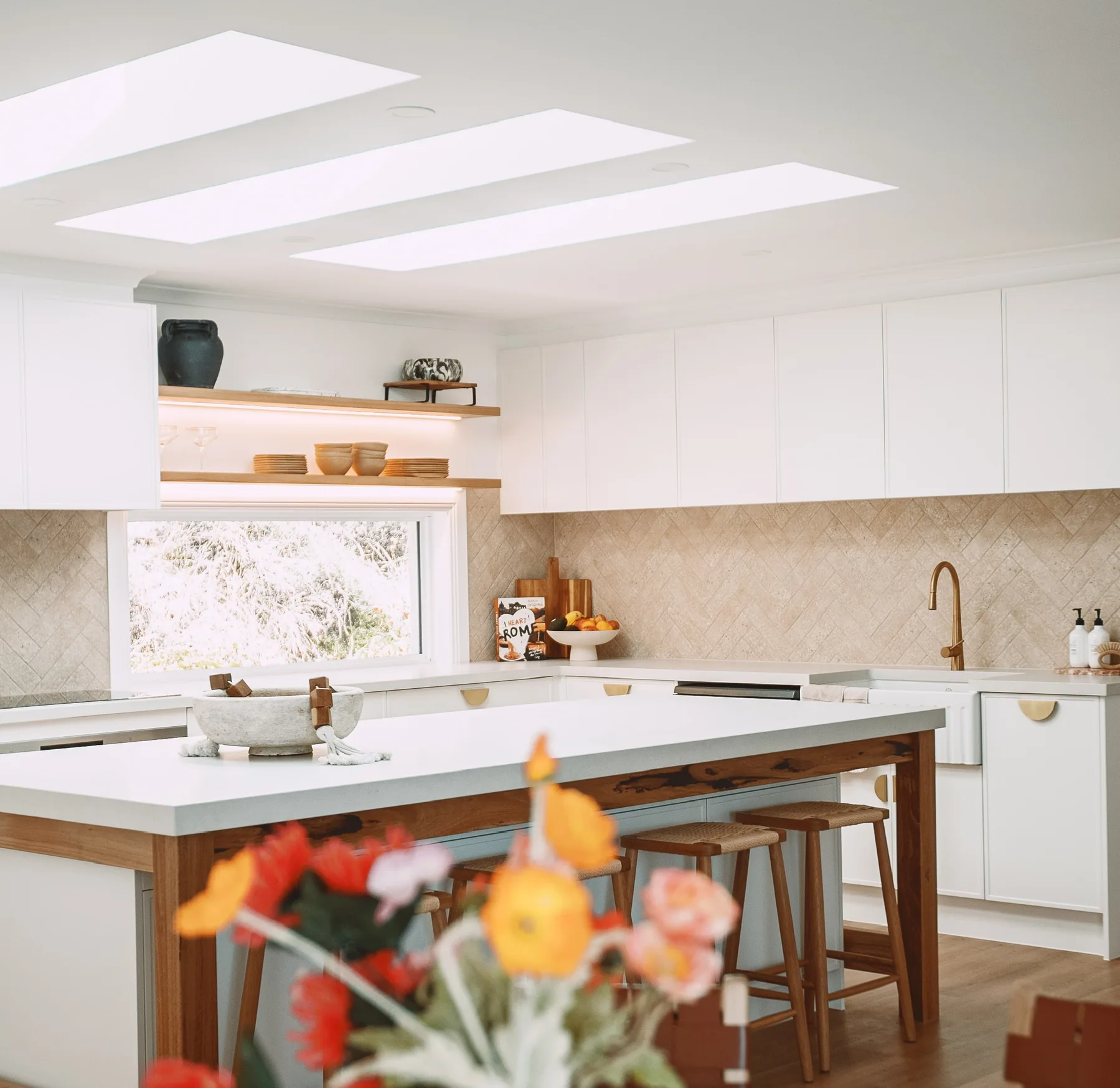 Kitchen with white flat-panel overhead cabinets, a white-topped island on a timber frame, timber stools, floating timber shelves and a herringbone tiled splashback.