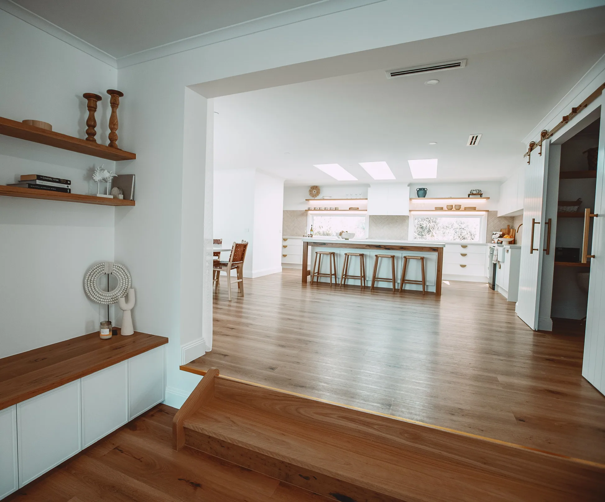 Built-in bench seat with a timber top and white doors under floating timber shelves, a timber step into the open-plan kitchen, and sliding white barn doors with brass handles on the right.