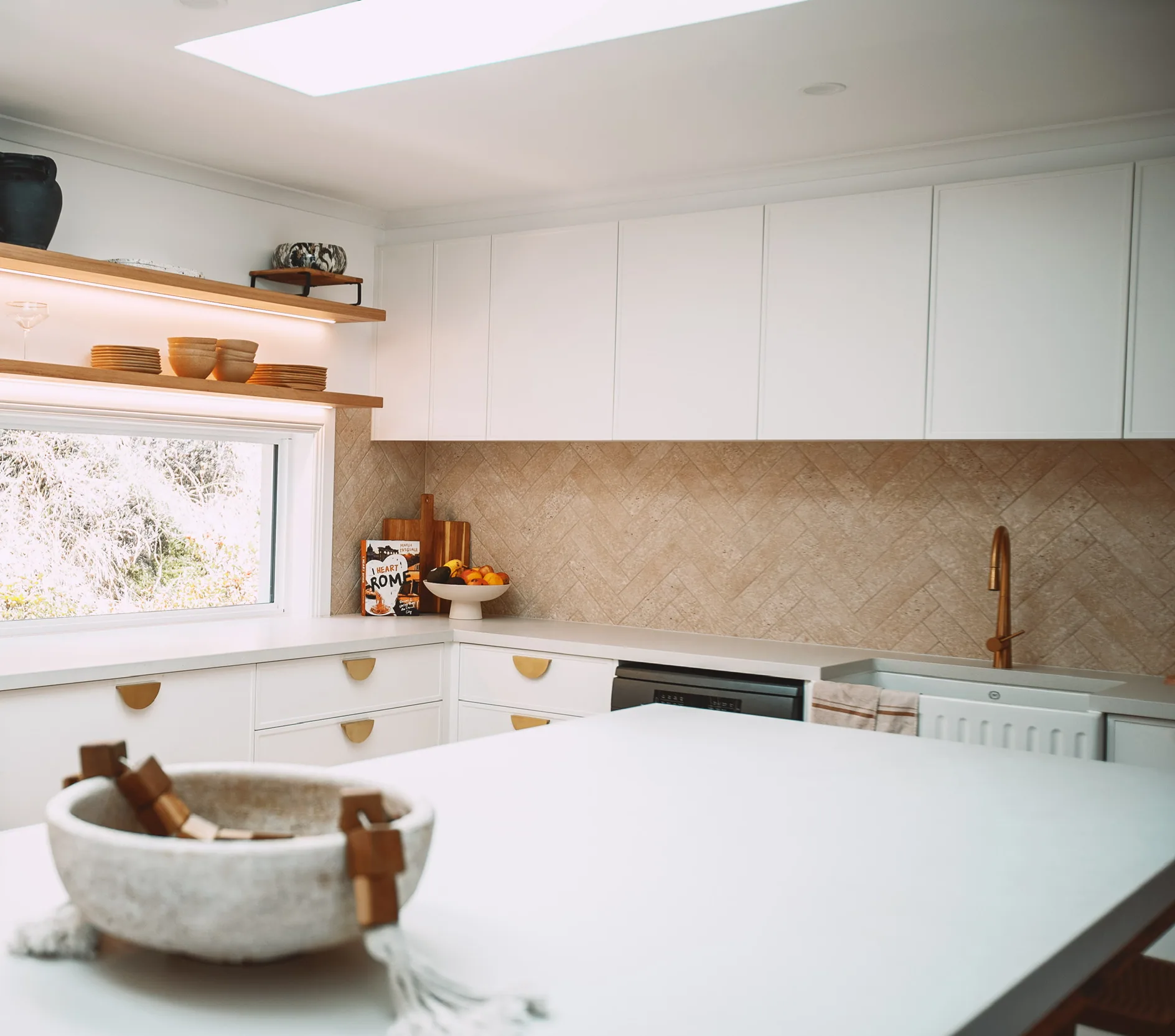 Corner of a kitchen with white flat-panel overheads, white drawers with brass half-moon pulls, lit floating timber shelves, a white butler sink and a white-topped island in the foreground.