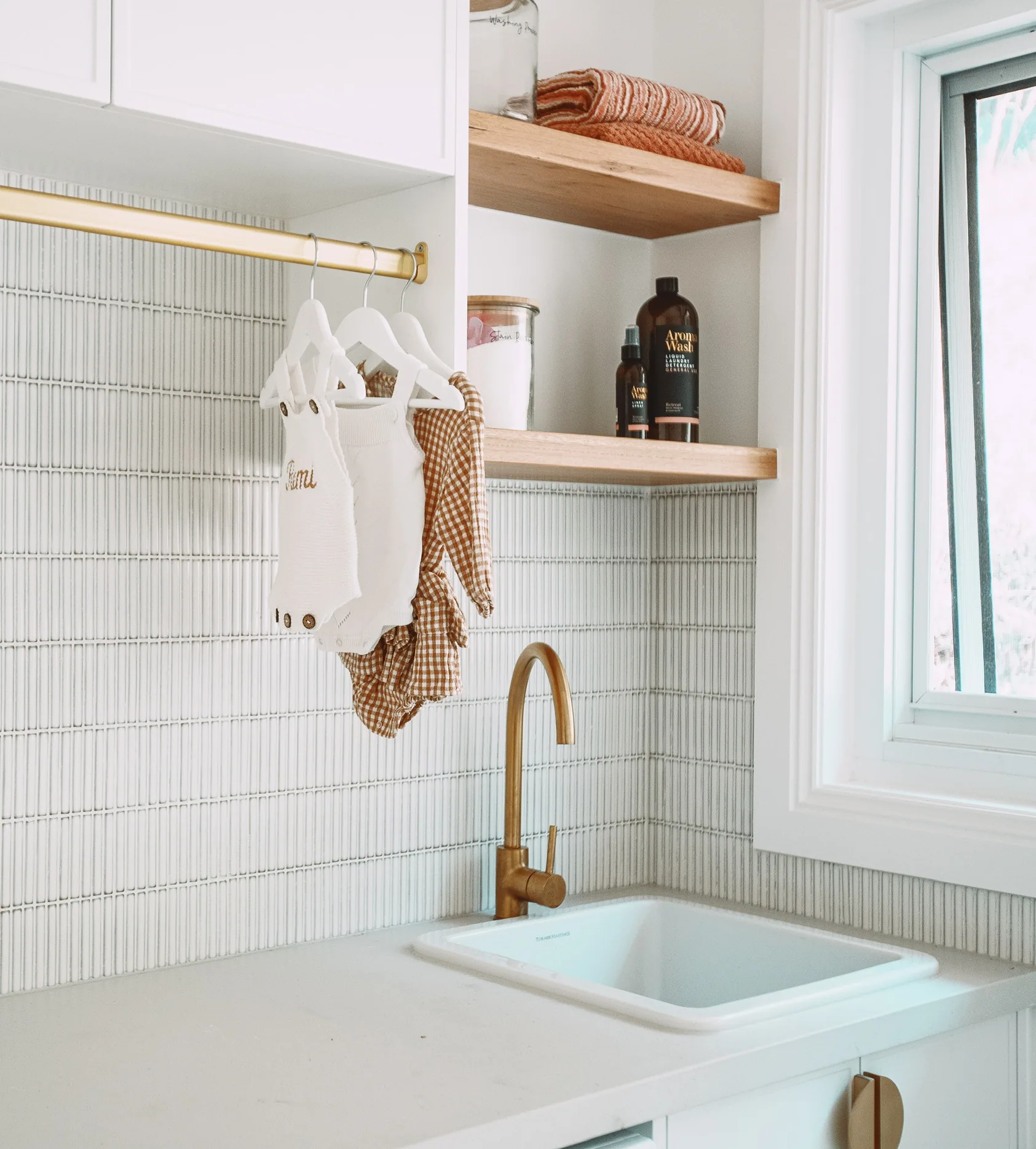 Laundry with white overhead cabinets, a brass hanging rail, floating timber shelves, white finger-tile splashback, brass gooseneck tap and inset sink.
